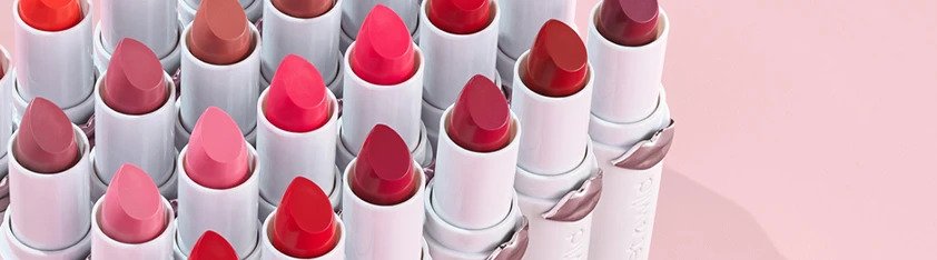 Rows of lipsticks in various shades of pink, red, nude, and coral in white tubes with metallic lip imprints on a soft pink background