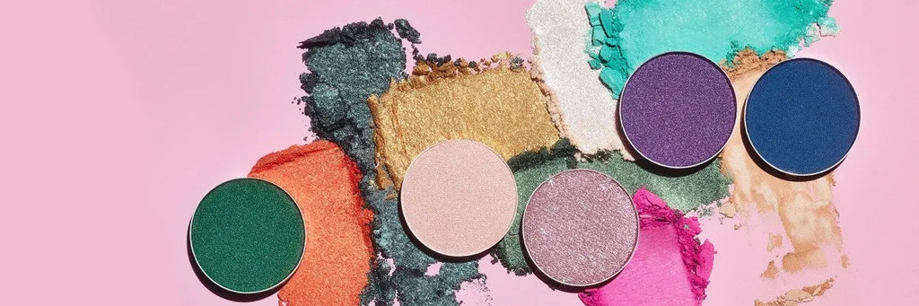 Crushed and pressed colorful eyeshadows in various shades including green, orange, gold, lilac, pink, purple, and blue scattered on a pink background