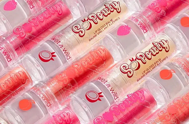 Multiple tubes of Wet n Wild So Pouty Lip Gloss Balm arranged diagonally, showcasing various fruity flavors and colorful packaging with icons like strawberries, coconuts, and peaches.